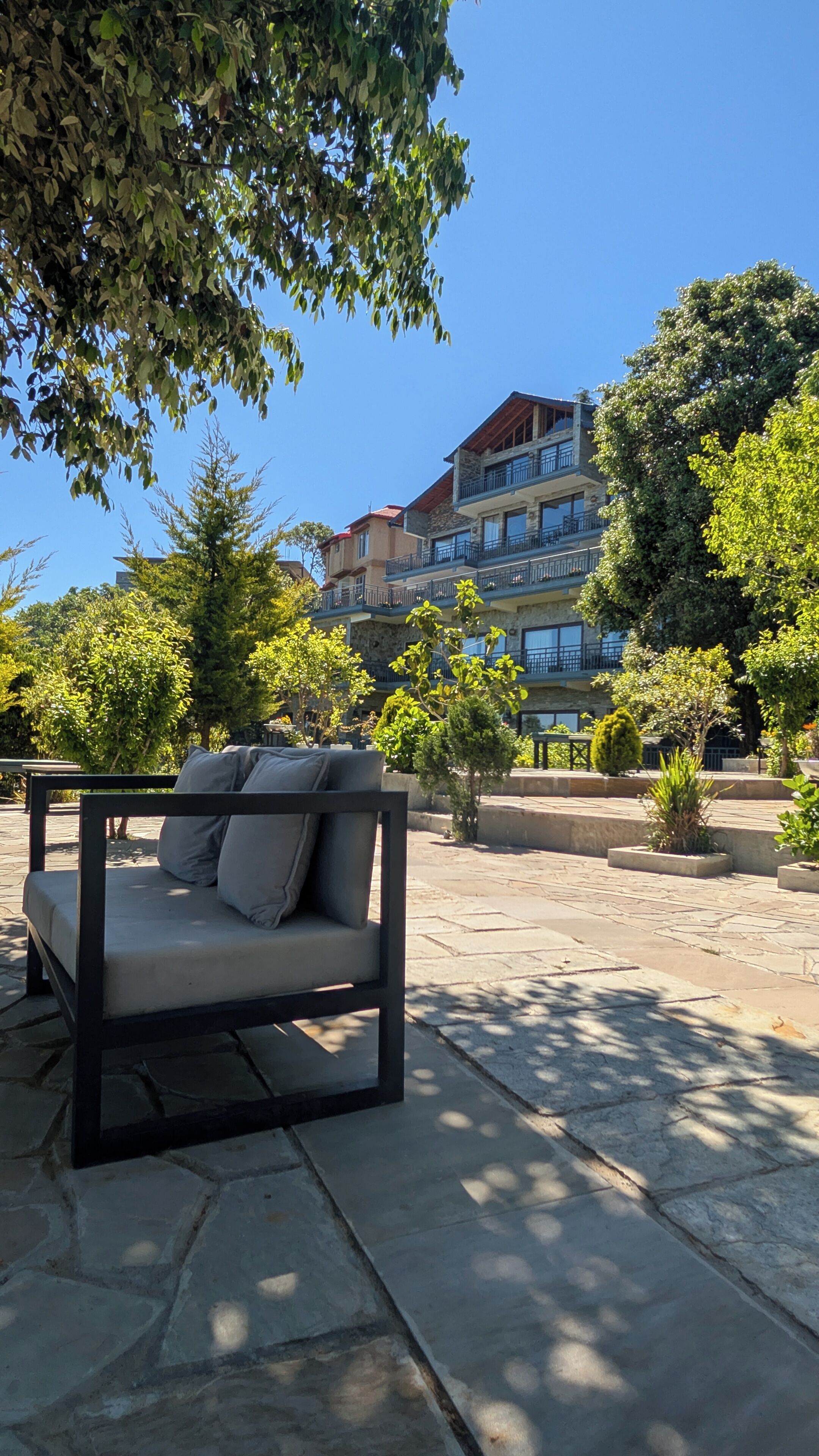 An outdoor seating area with a sofa and a table, surrounded by trees and a building with multiple balconies.