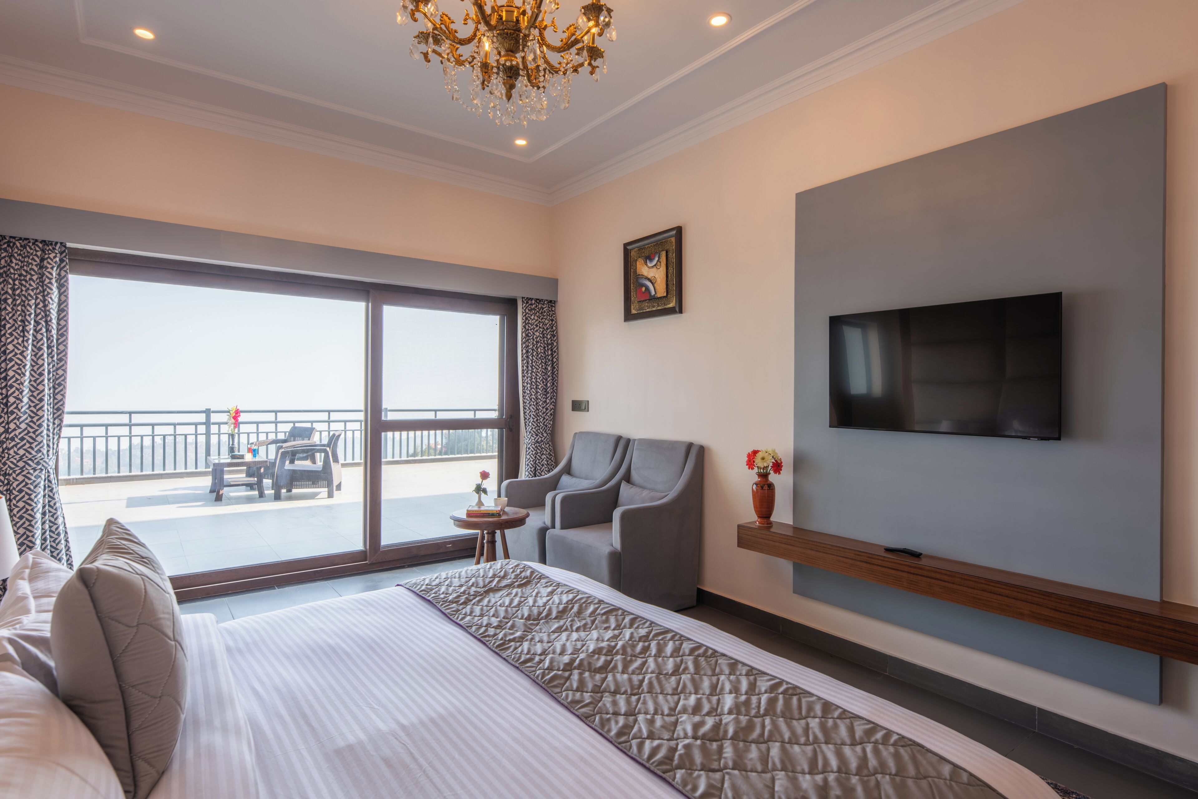 Habitación de hotel con una cama grande, un televisor montado en la pared, una silla, una mesita con un florero y un balcón con barandilla y una mesa.