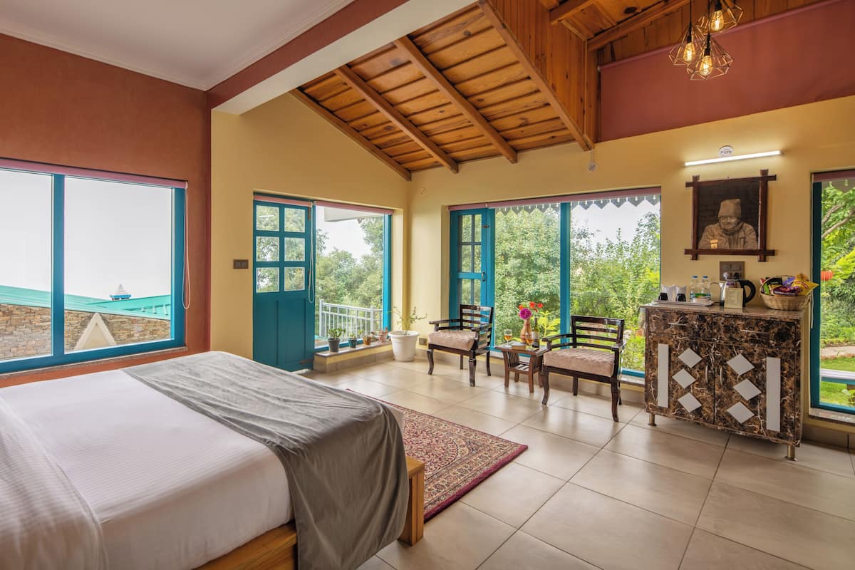 A bedroom with a bed, a desk, chairs, a view of the sea, and a balcony with potted plants.