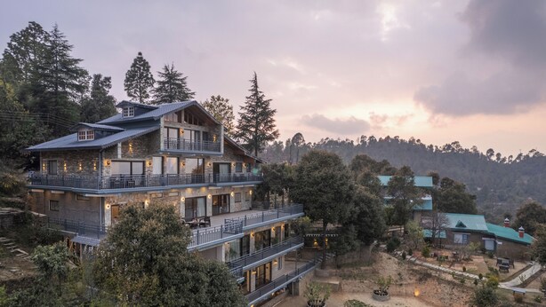 A multi-story building with balconies, surrounded by trees and a hilly landscape.
