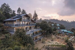 A multi-story building with balconies, surrounded by trees and a hilly landscape. - Dharohar Retreat Mukteshwar (Kosiyakutoli)