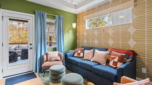 Sala de estar com um sofá azul, uma poltrona rosa e pufes. Há uma porta com cortinas azuis, uma janela com vista para árvores e uma parede estampada.
