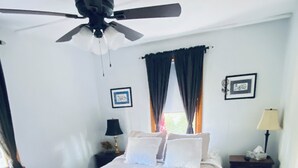 Ein Schlafzimmer mit Bett, Deckenventilator, Fenster mit Vorhängen, Nachttisch mit Lampe und einem Wandbild.