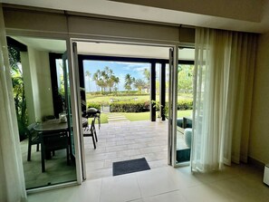 Ein modernes Zimmer mit einer Schiebeglastür, die auf eine Terrasse mit Grill, Gartenmöbeln und Blick auf einen gepflegten Garten und Palmen führt.