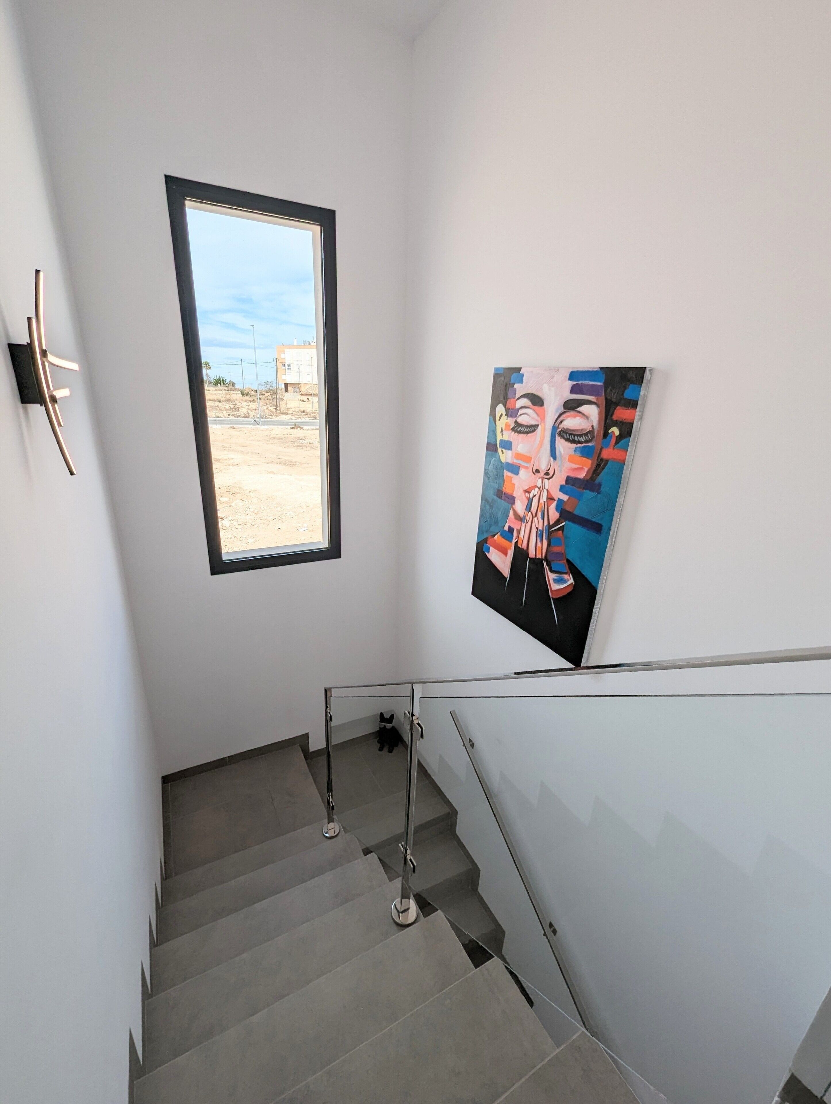 Un escalier avec une rampe en verre, une fenêtre donnant sur un immeuble et un tableau accroché au mur.