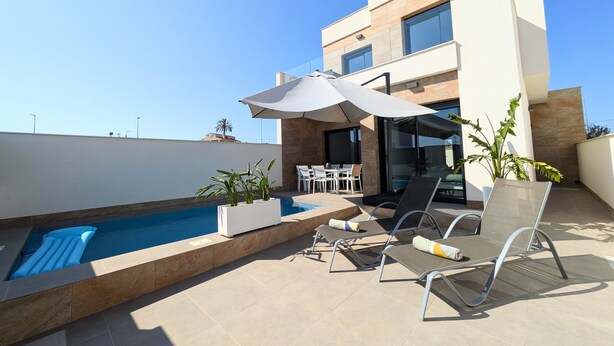 A modern outdoor pool area with lounge chairs and a table, surrounded by a building with large windows and a clear sky.