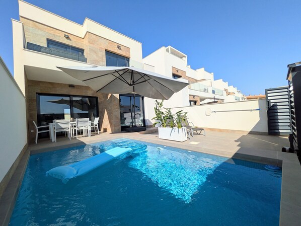 A modern outdoor pool area with a clear blue pool, white lounge chairs, and a large umbrella.