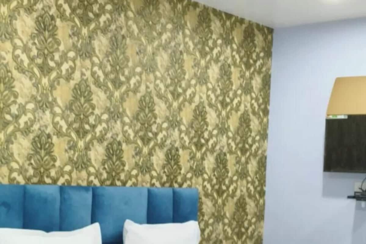 Una habitación de hotel con una cama, un cabecero azul, papel tapiz estampado, un ventilador de techo y un televisor montado.