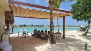 Uma área de estar coberta ao ar livre, com mesas e cadeiras, uma palmeira e vista para a praia e o oceano.