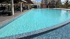 Piscina azul translúcida com borda em azulejo, área de estar coberta e edifícios modernos ao fundo.