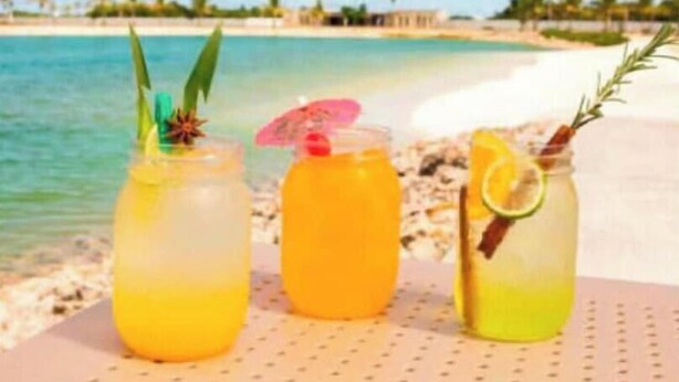 Três bebidas sobre uma mesa, com uma praia e oceano ao fundo.