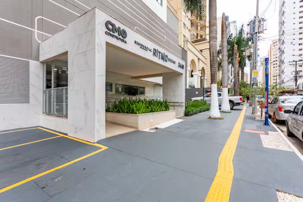 Un edificio moderno con un cartel de CMO Construcciones, un complejo residencial llamado RITMO BUENO, y un estacionamiento delimitado con líneas amarillas.