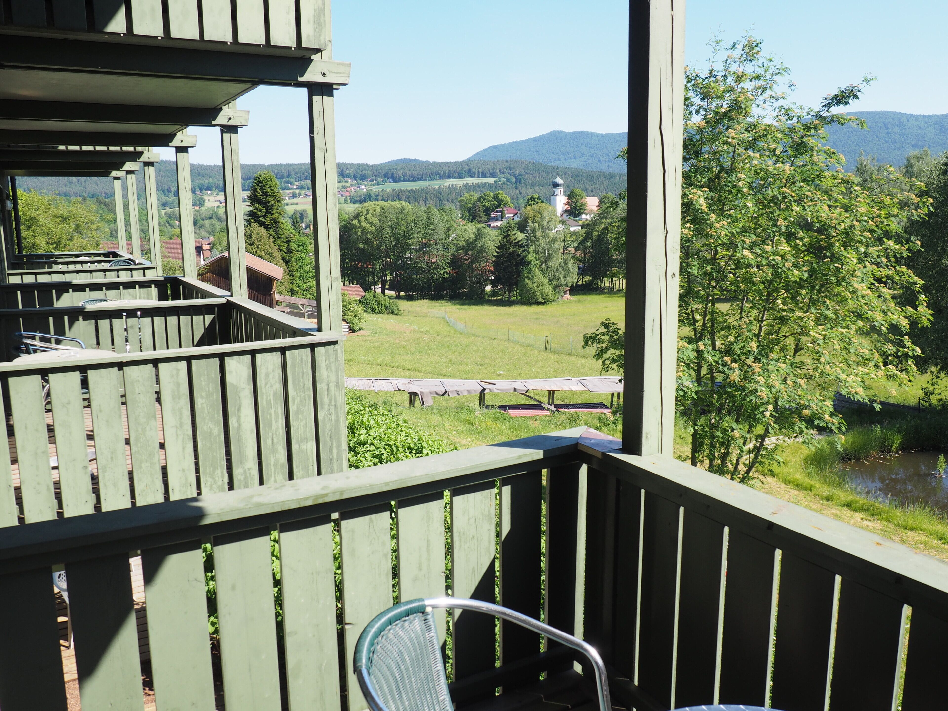 A wooden balcony with a view of a green landscape, a bench, and a chair.