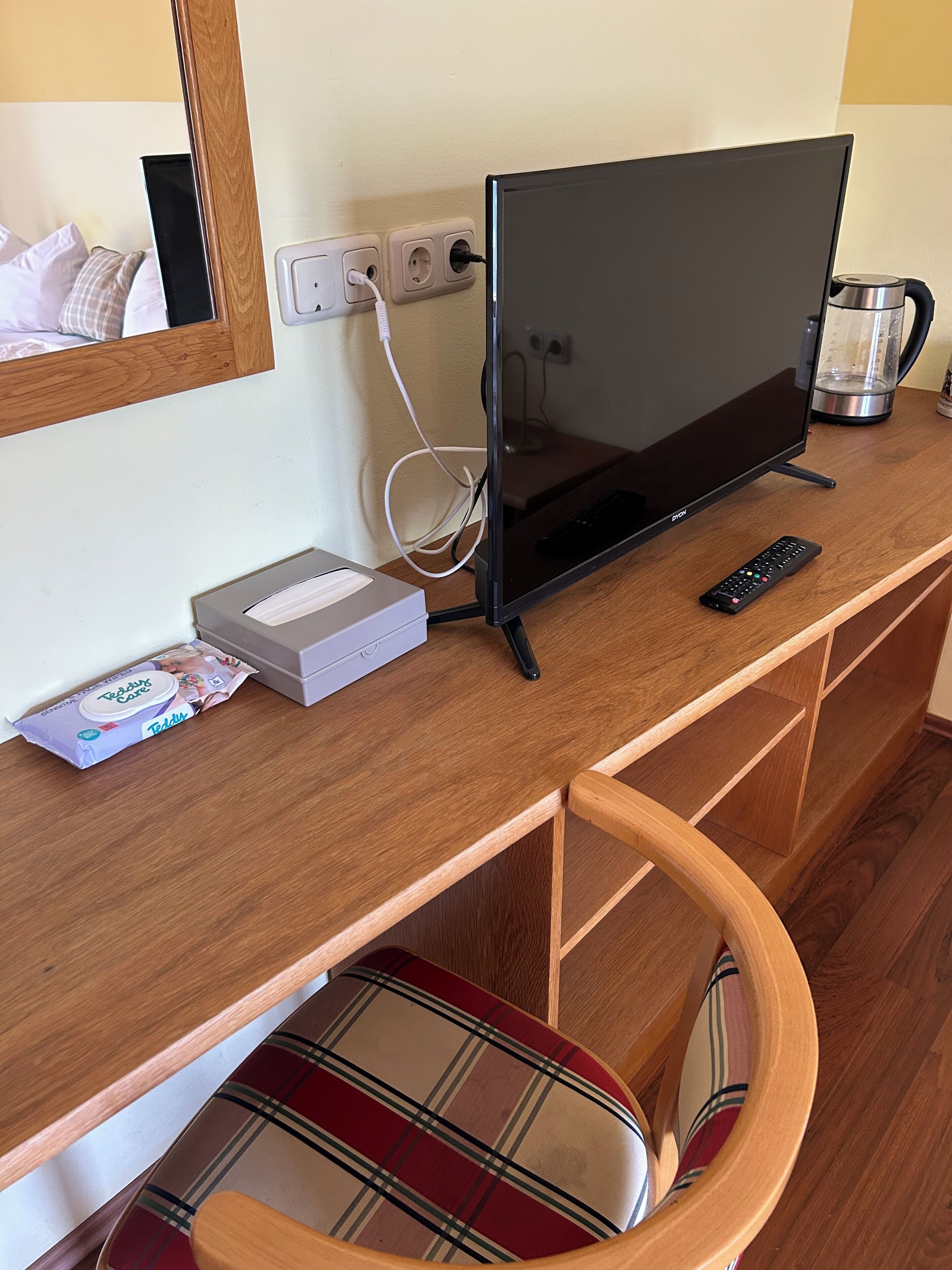 A wooden desk with a flat-screen TV, a remote control, a box of tissues, and a kettle.