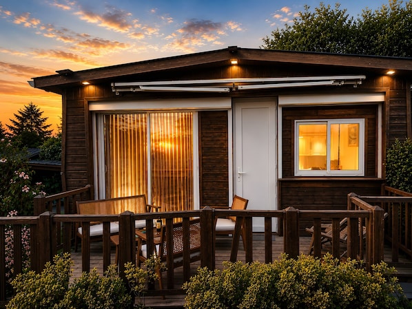 Une cabane en bois dotĂ©e dâune porte-fenĂȘtre coulissante, dâune terrasse avec du mobilier et dâune fenĂȘtre avec un Ă©clairage intĂ©rieur.