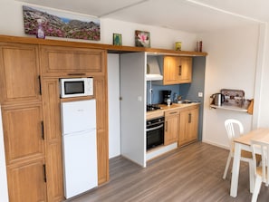 Une cuisine compacte dotĂ©e de meubles en bois, dâun four Ă micro-ondes encastrĂ©, dâun rĂ©frigĂ©rateur, dâune cuisiniĂšre et dâune machine Ă cafĂ©.
