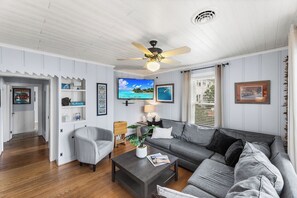 Ein Wohnzimmer mit grauem Sofa, weißem Sessel, Couchtisch aus Holz und Deckenventilator.