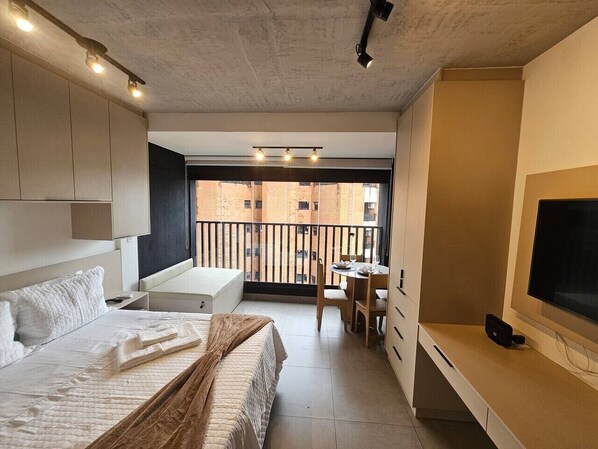 A modern bedroom with a bed, bedside table, and a TV. There is a balcony with a view of buildings. - Studio Aconchegante - Brooklin (São Paulo)