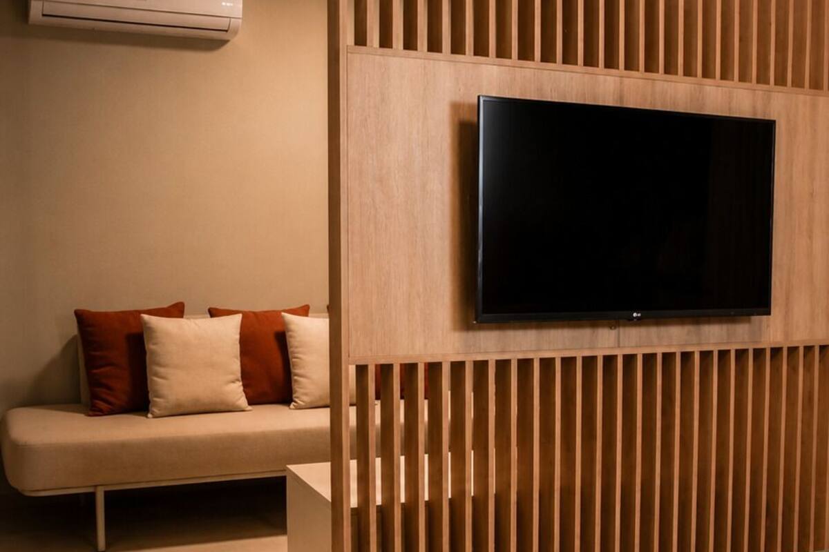 A bedroom with a bed, a TV mounted on a wooden panel, a sofa, and a wall-mounted air conditioner.