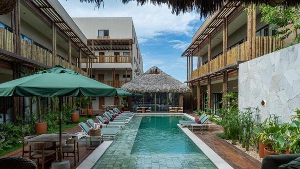 Un área de piscina con sillones de descanso y sombrillas, rodeada de edificios con balcones de madera y una estructura de techo de paja.