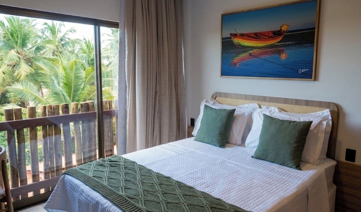 A bedroom with a bed, pillows, a painting, and a view of palm trees through a window.