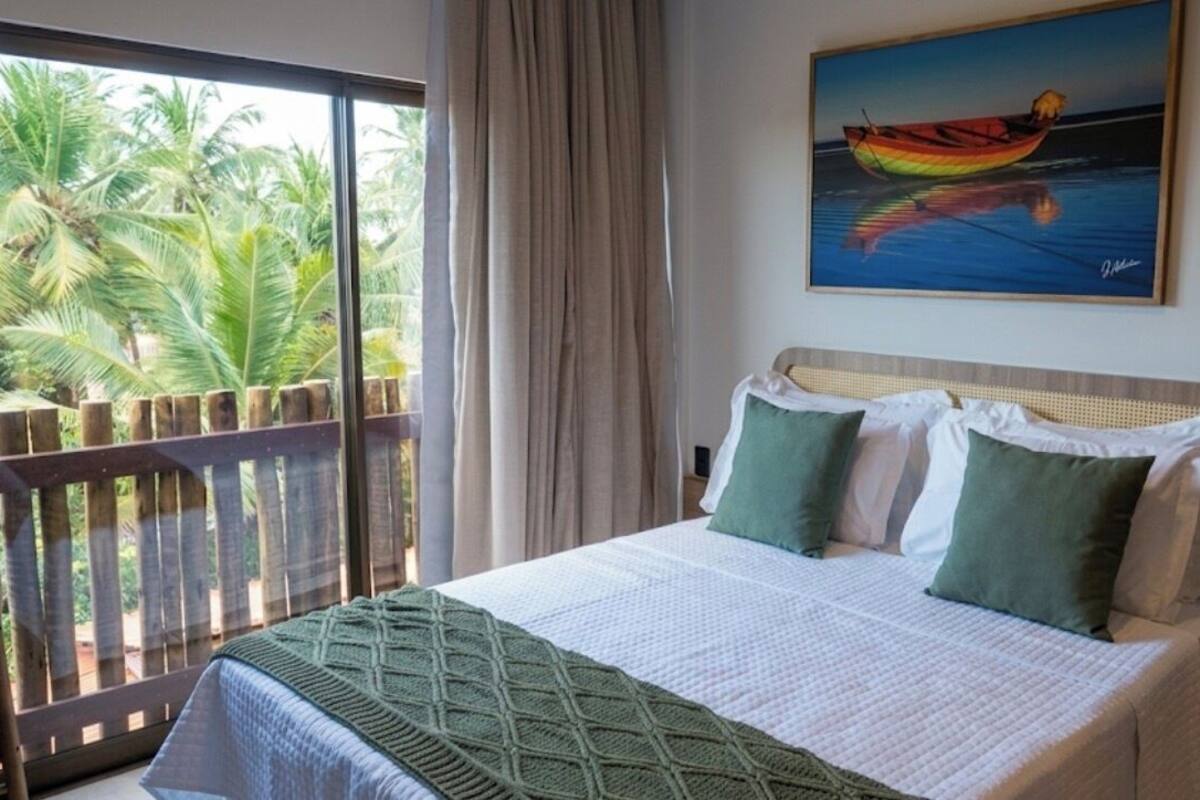 A bedroom with a bed, pillows, a painting, and a view of palm trees through a window.