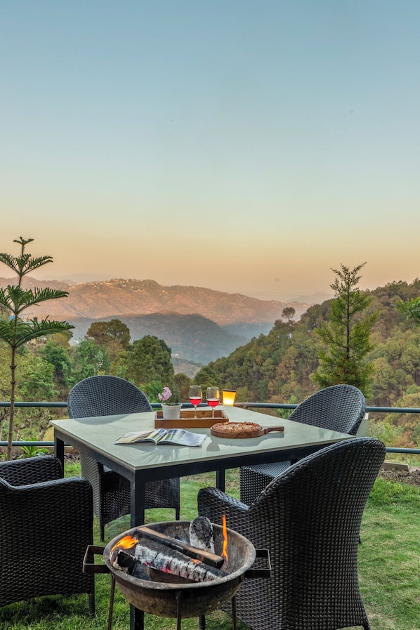 A dining table with chairs, a fire pit, and a scenic view of hills. - Alaya Stays Live Life 2BHK Apartment (Kasauli)