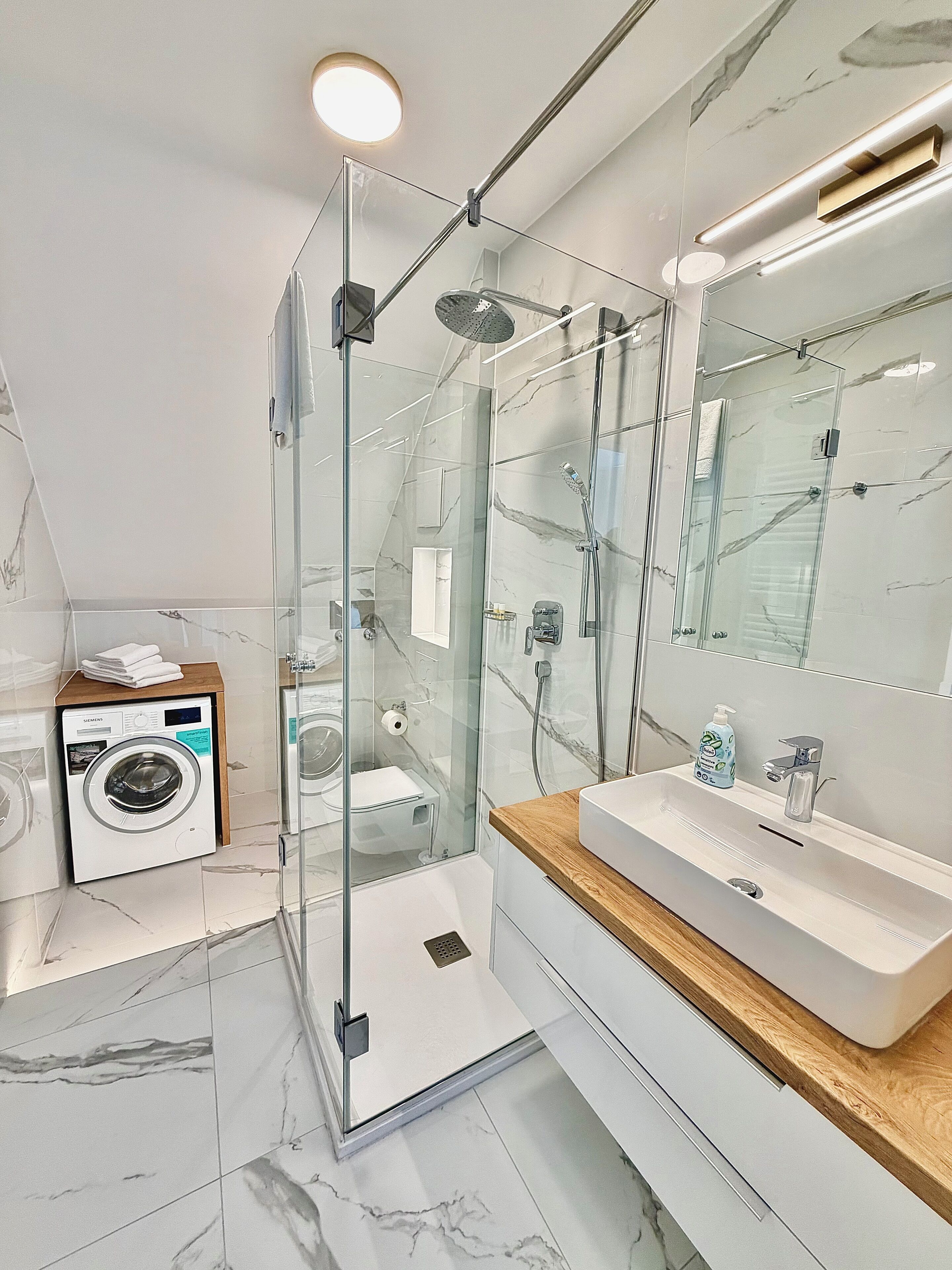Une salle de bain moderne dotée d’une douche à parois de verre, d’un lave-linge encastré et d’un meuble-lavabo avec miroir.