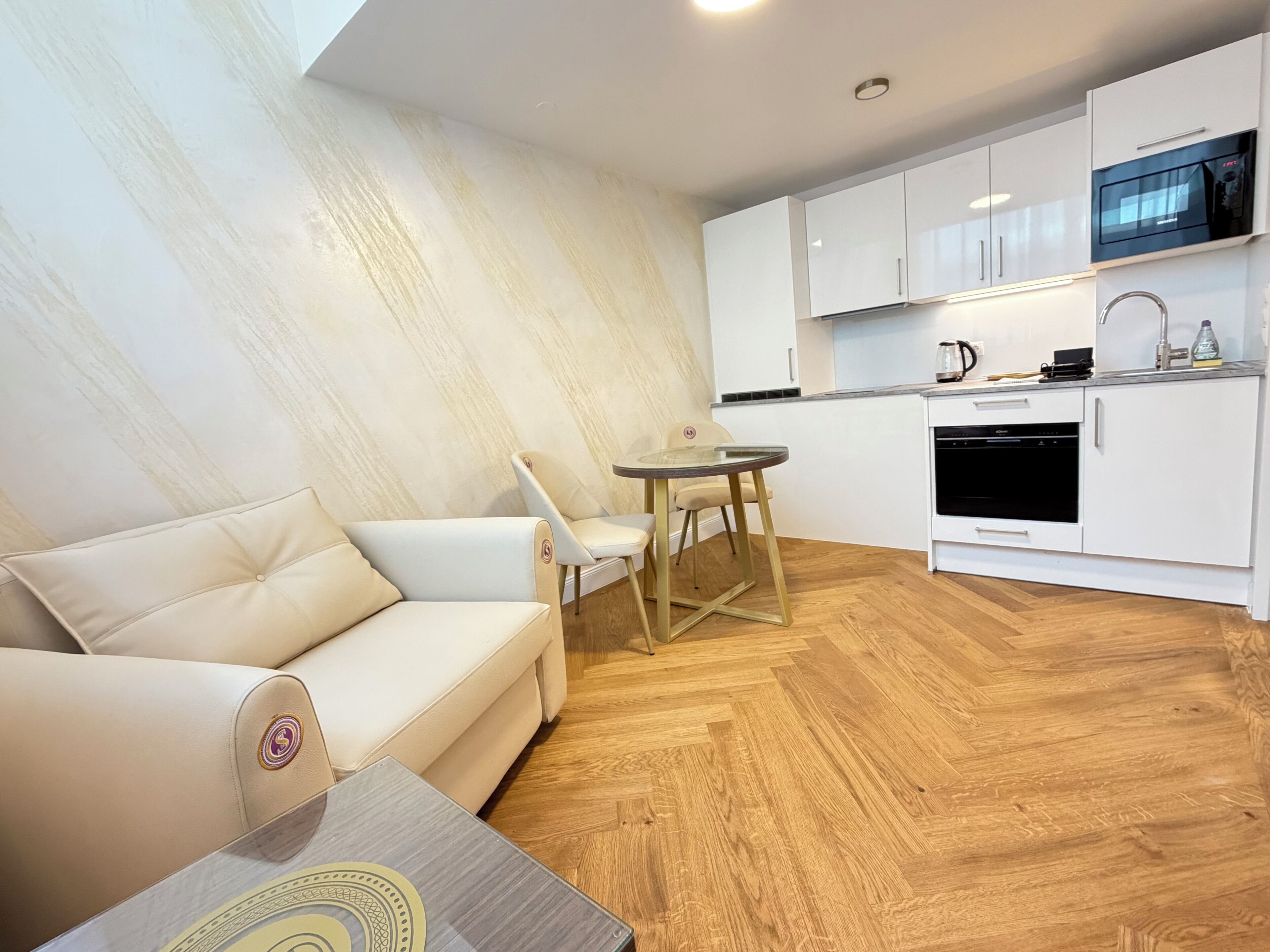 A modern kitchen with white cabinets, a microwave, and a coffee maker. There is a beige sofa and a small round table with chairs.