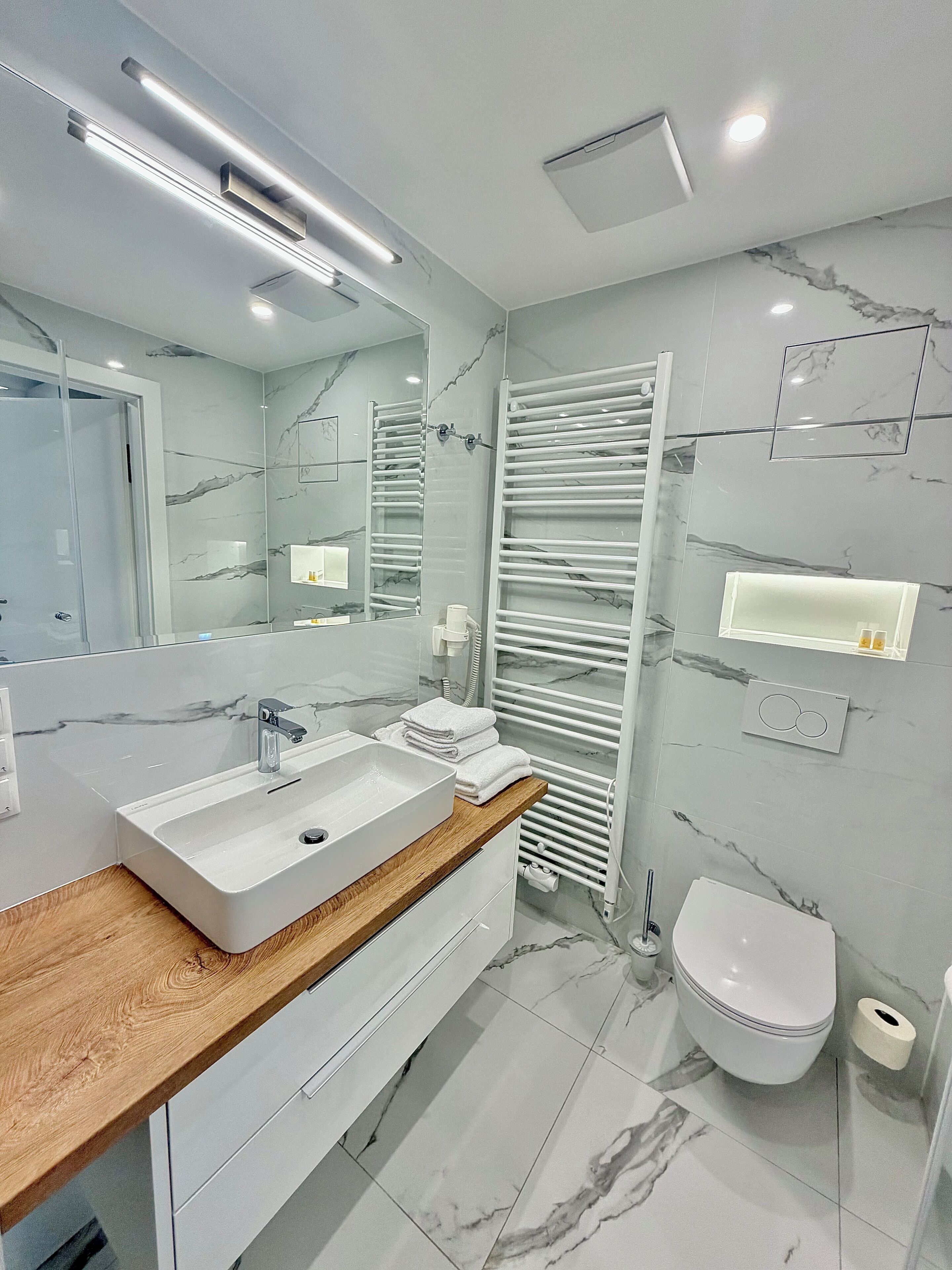 Un baño moderno con tocador de madera, un espejo grande, un lavabo blanco, un inodoro y un perchero para toallas.
