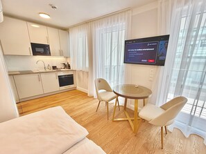 Ein modernes Hotelzimmer mit Küchenzeile, einem Essbereich mit Glastisch und einem an der Wand montierten Fernseher.