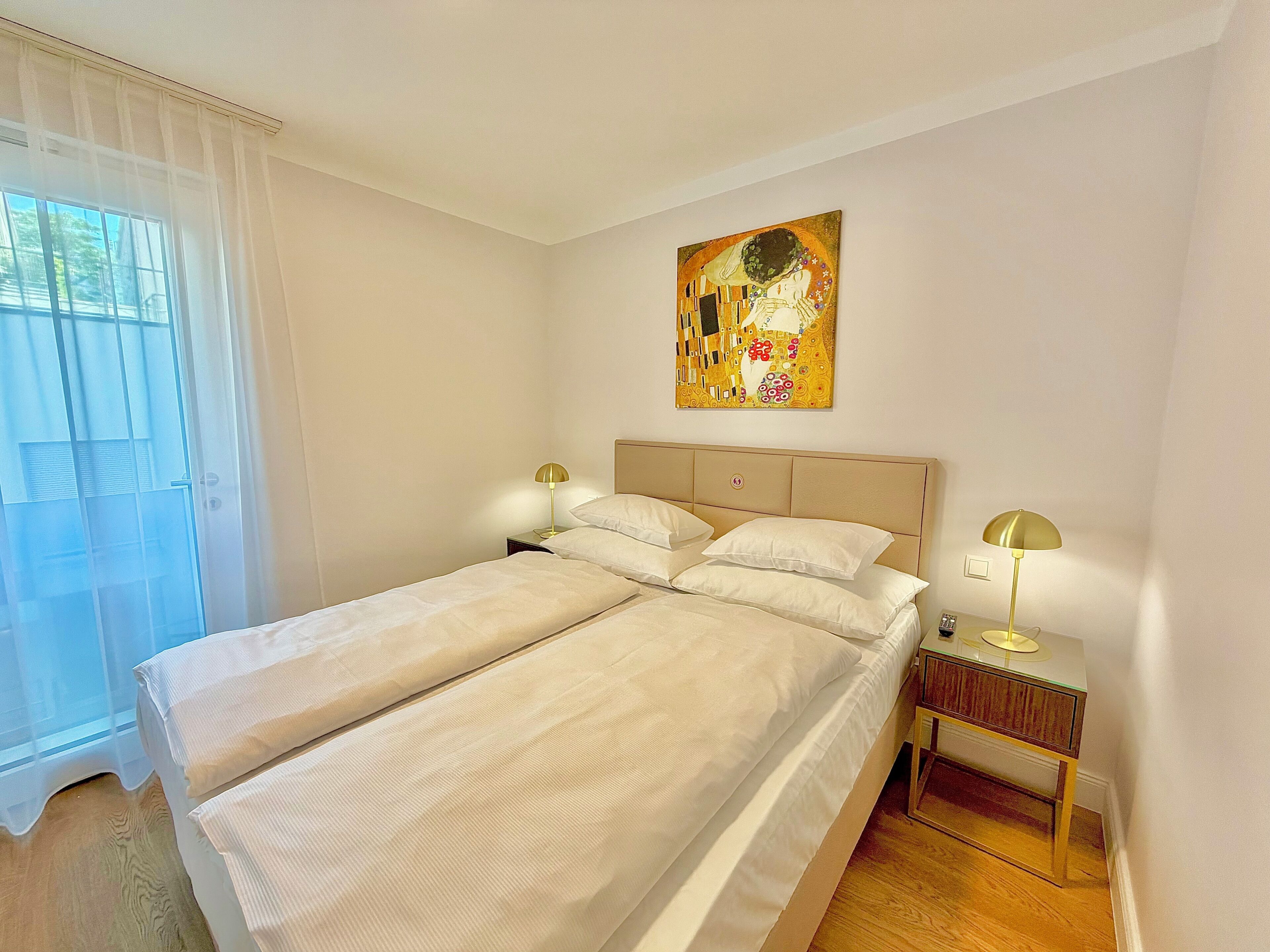 A double bed with two bedside tables, a painting on the wall, and a window with sheer curtains.
