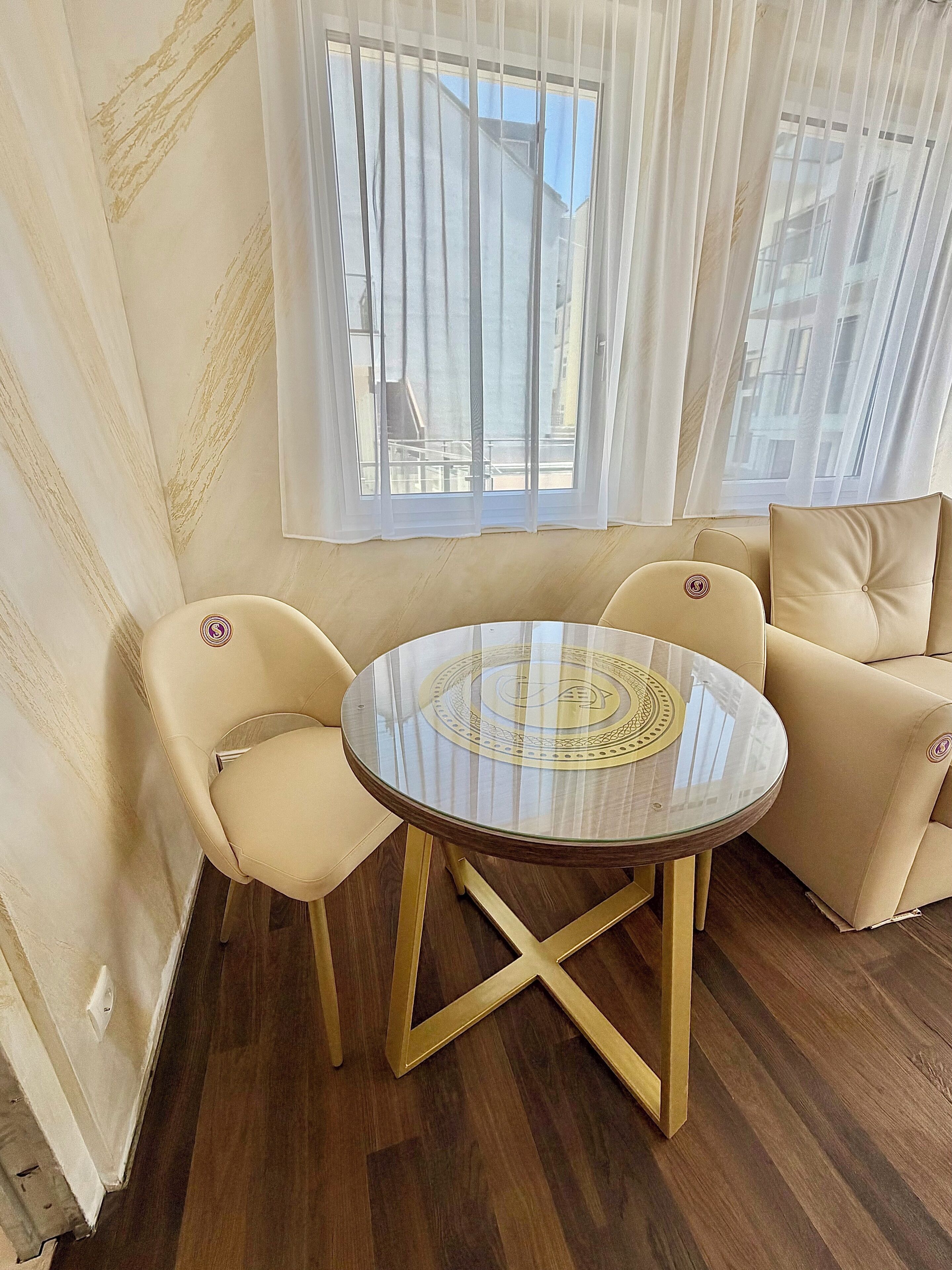 A room with a round glass table, beige chairs, a sofa, and a window with curtains.