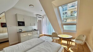 A modern hotel room with a bed, kitchenette, dining area, and a view of the city. - Business Quartier Belvedere (Vienna)