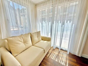 Ein Zimmer mit einem beigen Sofa, großen Fenstern mit durchsichtigen Vorhängen und Holzboden.