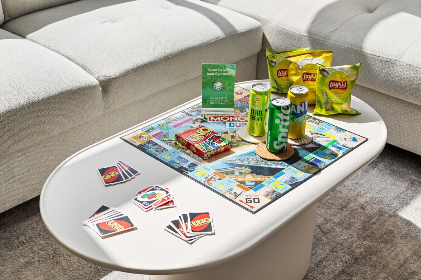 A board game setup on a white table with a Monopoly box, UNO cards, and soda cans.