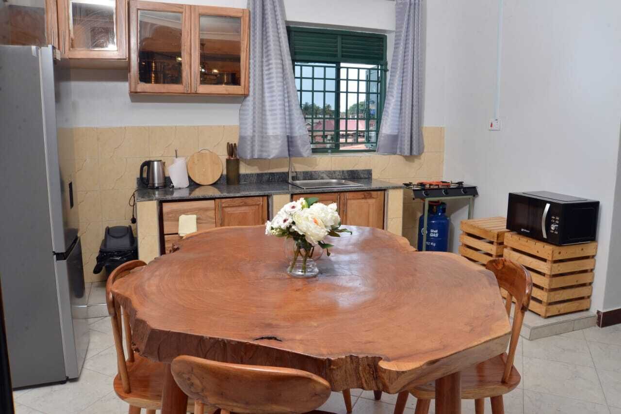 Una cocina con una mesa redonda de madera, sillas de madera, un refrigerador, un microondas y una ventana con cortinas.