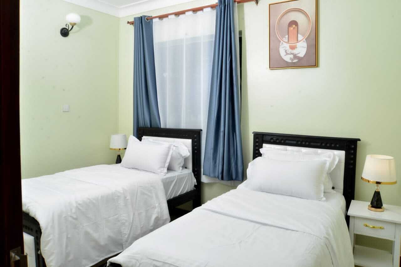 A hotel room with two single beds, a nightstand with a lamp, a window with blue curtains, and a framed picture on the wall.