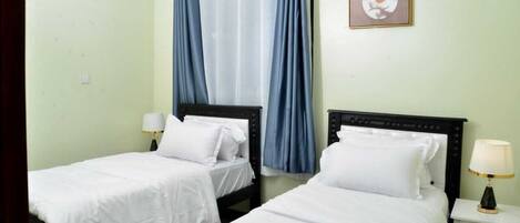 A hotel room with two single beds, a nightstand with a lamp, a window with blue curtains, and a framed picture on the wall.