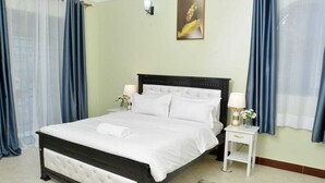 A neatly made bed with white linens, a bedside table with a lamp, and a painting on the wall. - Beautiful 2-bedroom corporate apartments within  Entebbe (Entebbe)