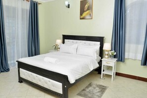A neatly made bed with white linens, a bedside table with a lamp, and a painting on the wall. - Beautiful 2-bedroom corporate apartments within  Entebbe (Entebbe)
