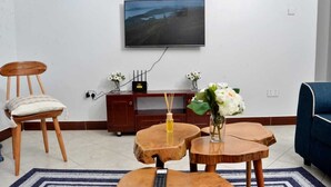 A living room with a TV mounted on the wall, a wooden coffee table with a remote control, a cushioned chair, and a blue sofa. - Beautiful 2-bedroom corporate apartments within  Entebbe (Entebbe)
