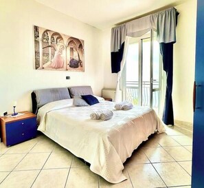 Une chambre à coucher avec un lit, une table de chevet, un tableau, une fenêtre avec des rideaux et un balcon.