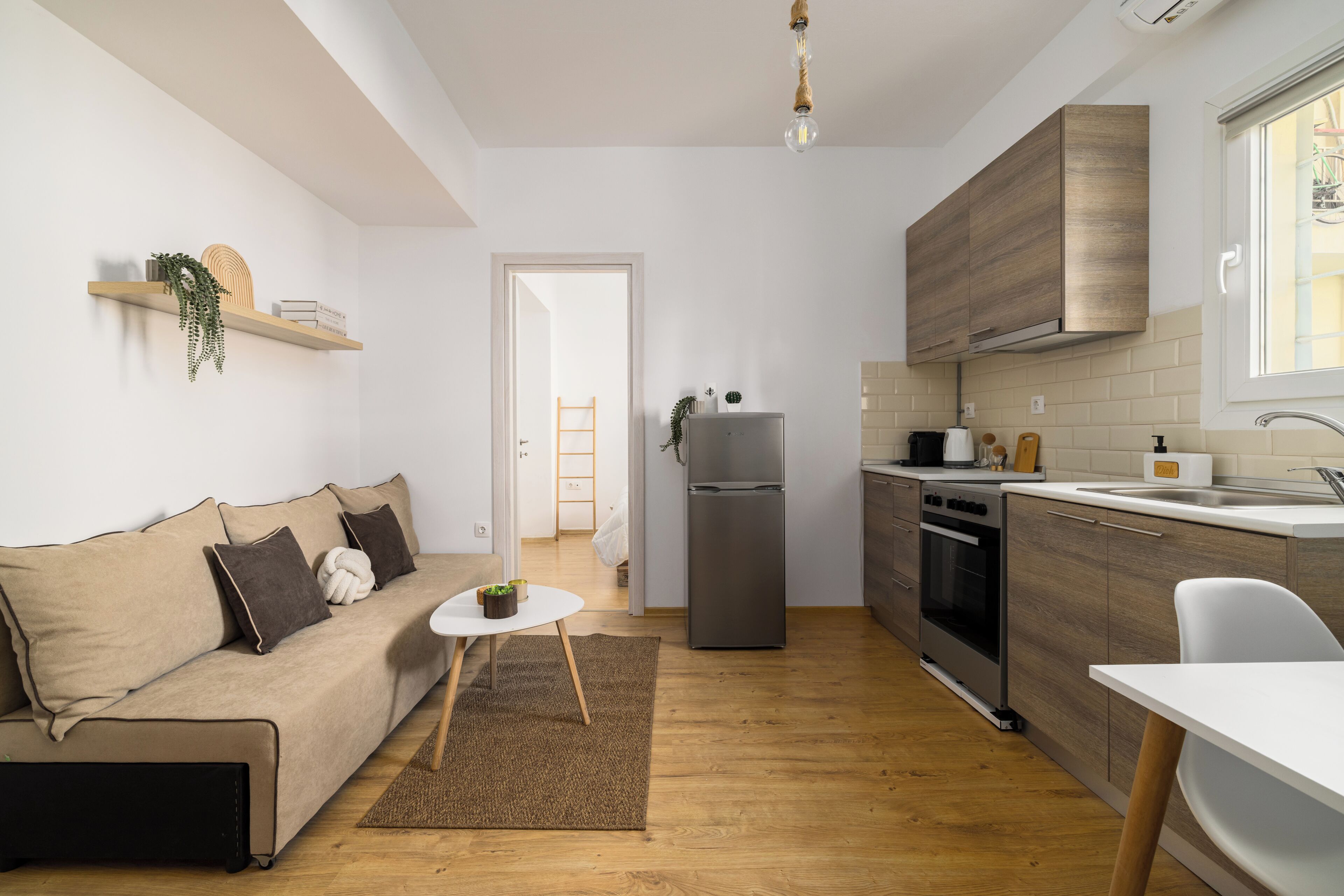 Cocina compacta con refrigerador, horno y lavamanos. Hay una mesa de comedor de madera y un sofá con cojines.
