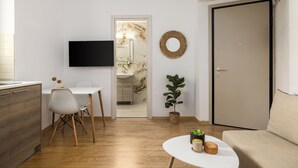 Un salon moderne avec un téléviseur à écran plat, un coin repas avec une table et des chaises blanches, un canapé, une plante en pot et une porte donnant accès à une salle de bain.