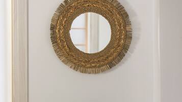 Een potplant met brede bladeren in een geweven mand, een geweven ronde spiegel aan de muur en een houten vloer.