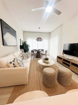 A modern living room with a sofa, coffee table, ottoman, TV, and dining area. - C5 -307 VISTA CANA / Panorama Lake (Punta Cana)