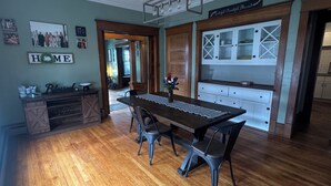 Un comedor con una mesa larga, sillas y una araña. Se observa una zona de cocina con armarios y un cartel sobre ellos.