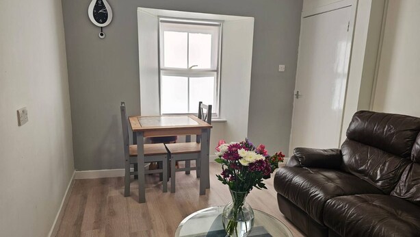 Una sala de estar con un sofá de cuero marrón, una mesa de centro de vidrio con un jarrón de flores, una mesa de comedor de madera con sillas y una ventana con cortinas blancas.