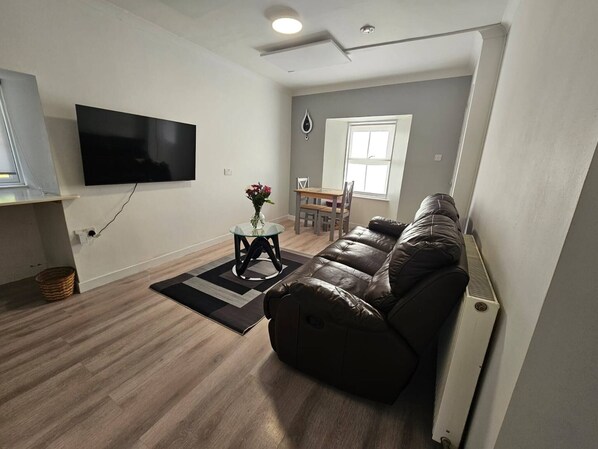 Ein modernes Wohnzimmer mit einer Ledersofa, einem flachbildfernseher an der Wand, einem kleinen runden Tisch mit Blumen in einer Vase und einem Essbereich mit Fenster.